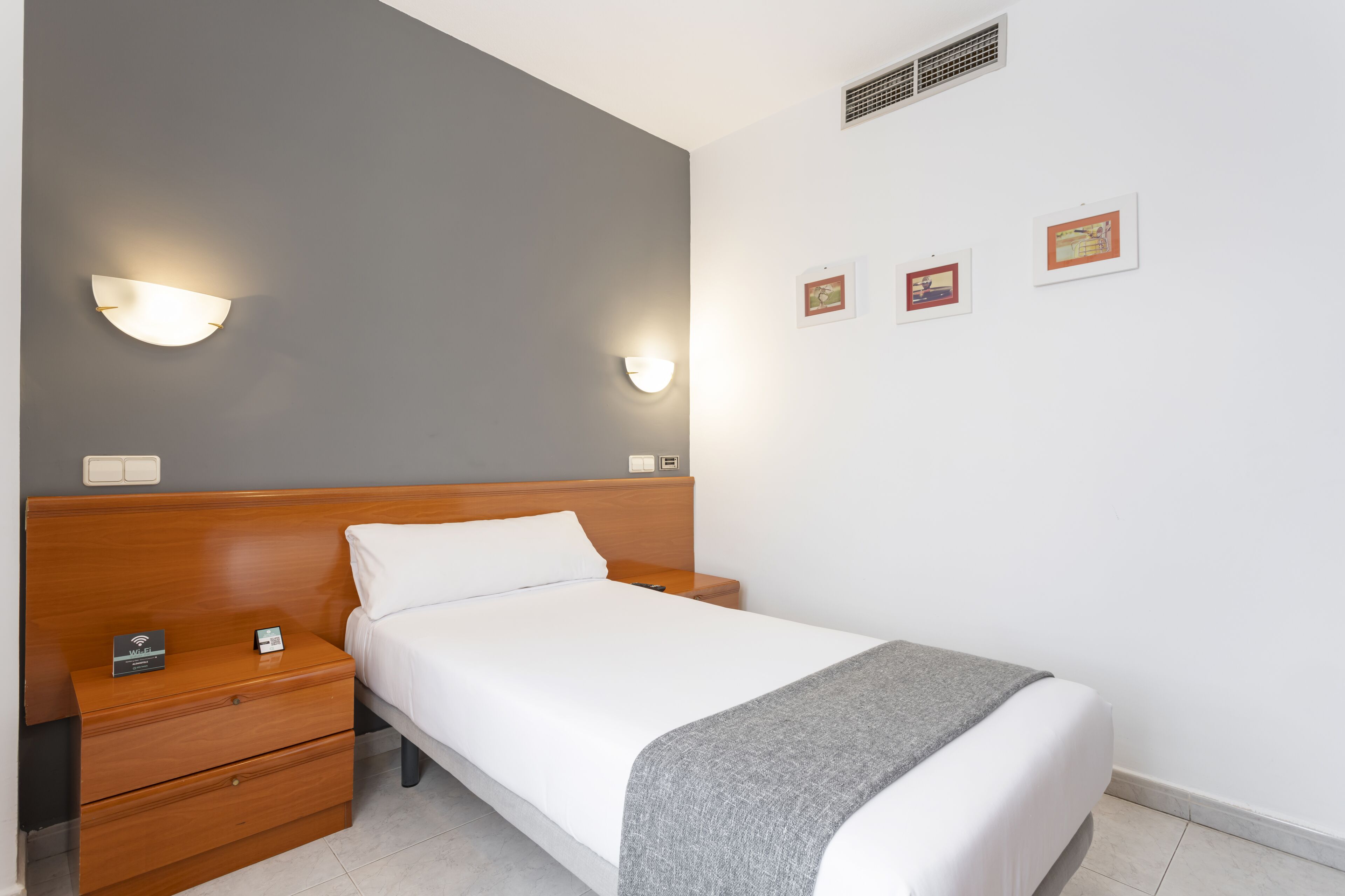 Una cama bien tendida con un edredón gris, una mesita de noche de madera con estación de carga y un aplique de pared.
