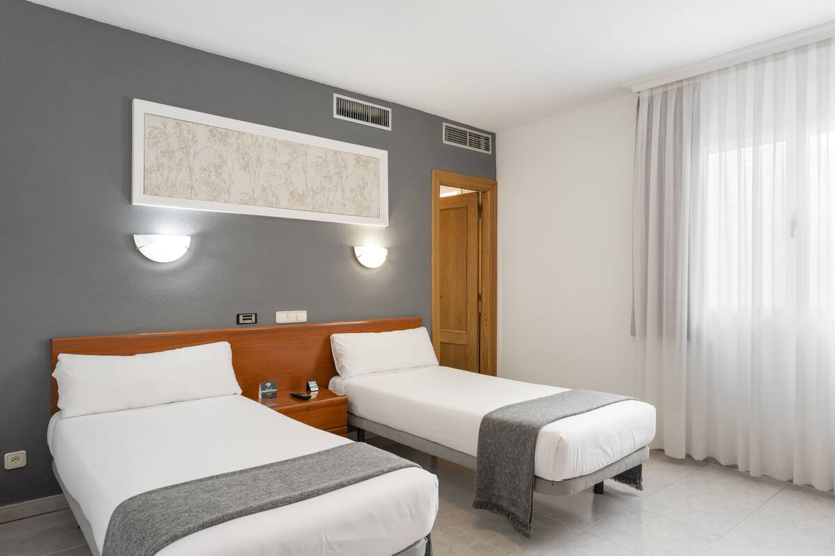 A hotel room with two beds, a wooden headboard, a wall-mounted light, a framed picture, and a window with sheer curtains.