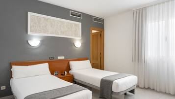 Habitación de hotel con dos camas, cabecero de madera, una lámpara de pared, un cuadro enmarcado y una ventana con cortinas traslúcidas.