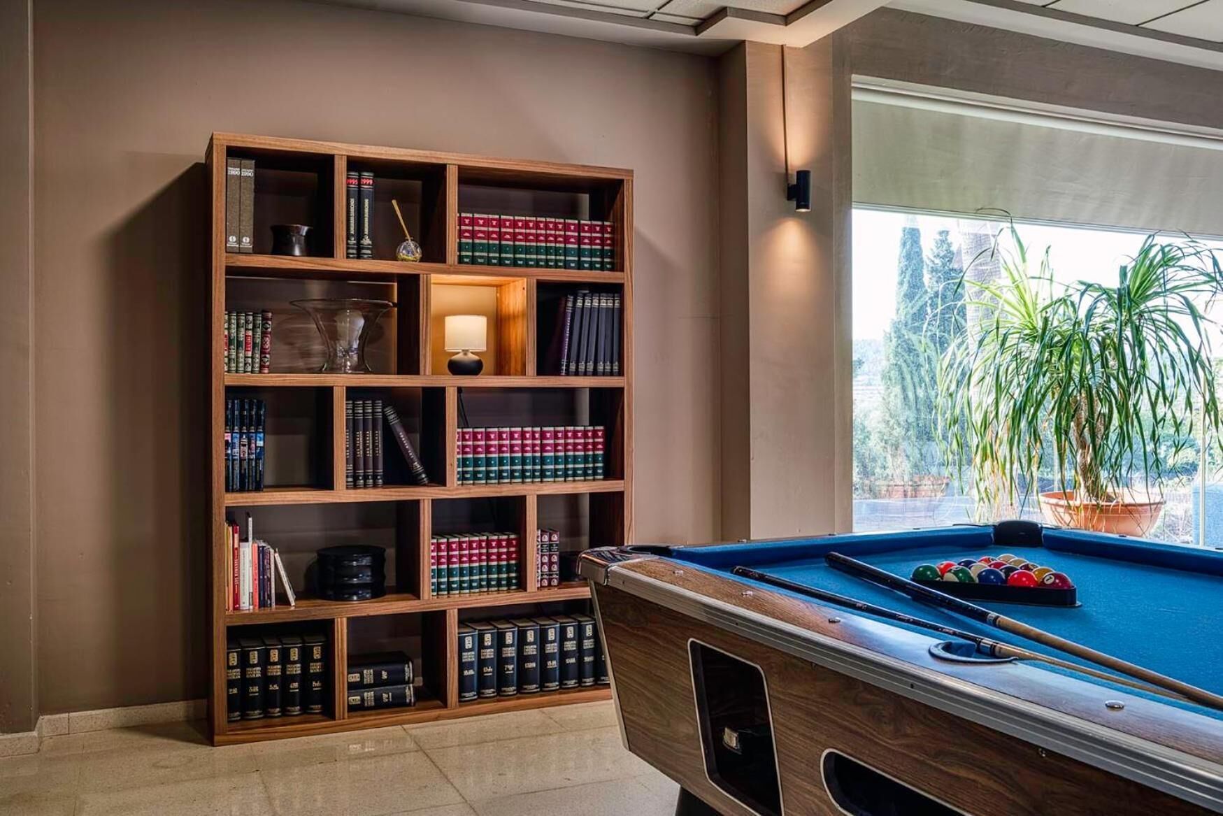 A pool table with a wooden frame, a bookshelf with books, and a window with a view of trees.