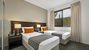 Habitación de hotel con dos camas, una mesita de noche con lámpara, una ventana con cortinas y vistas a una cerca de madera.