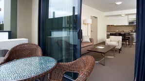 Una habitación de hotel moderna con una puerta de vidrio que da a un balcón, un área de comedor con mesa y sillas, y una kitchenette al fondo.