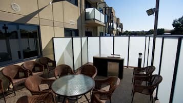 Een terras buiten met rieten meubels, een glazen tafel en een barbecue.