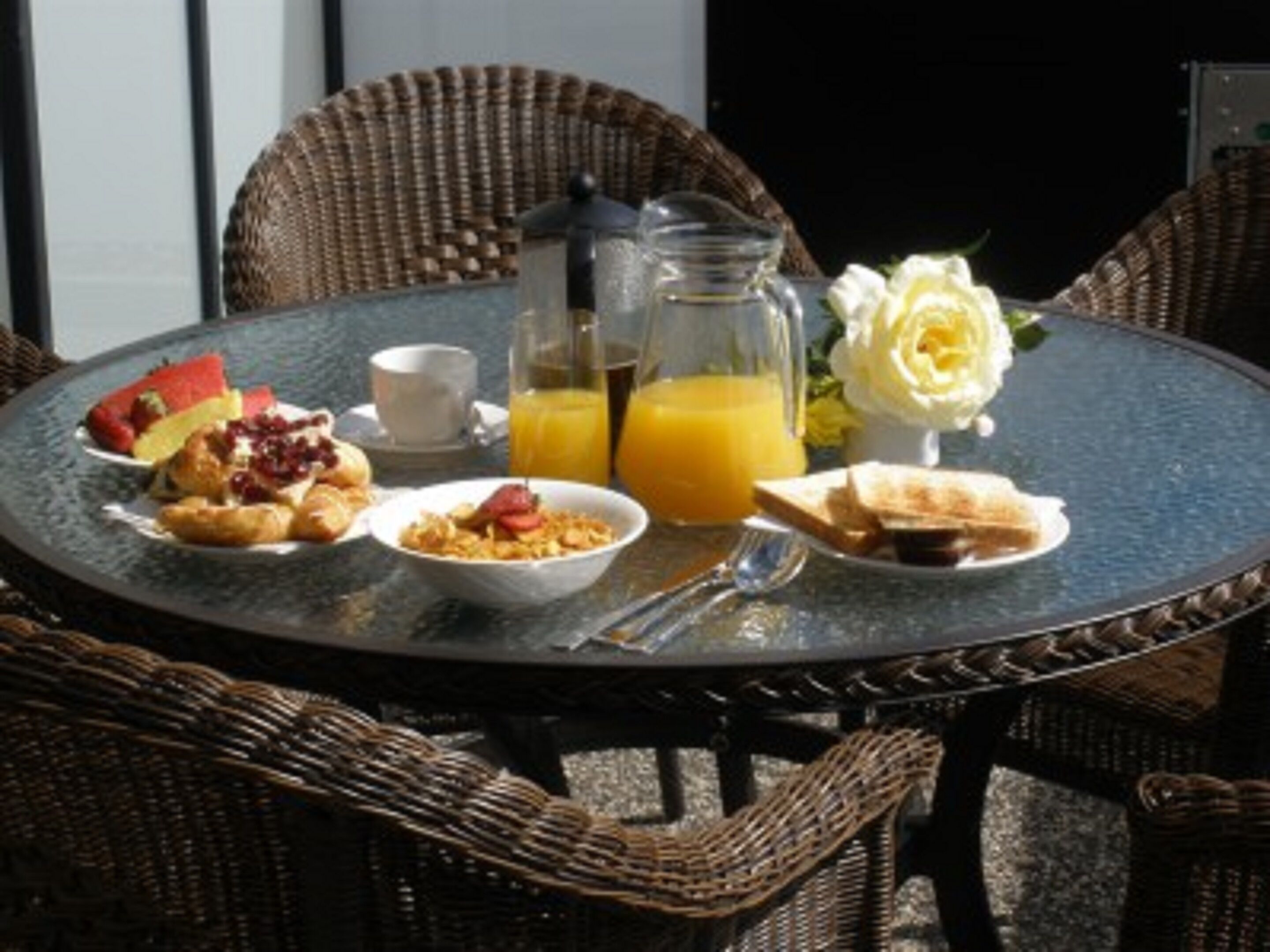 A round table with breakfast items including toast, fruit, and cereal, accompanied by a pitcher of orange juice and a vase with a single flower.