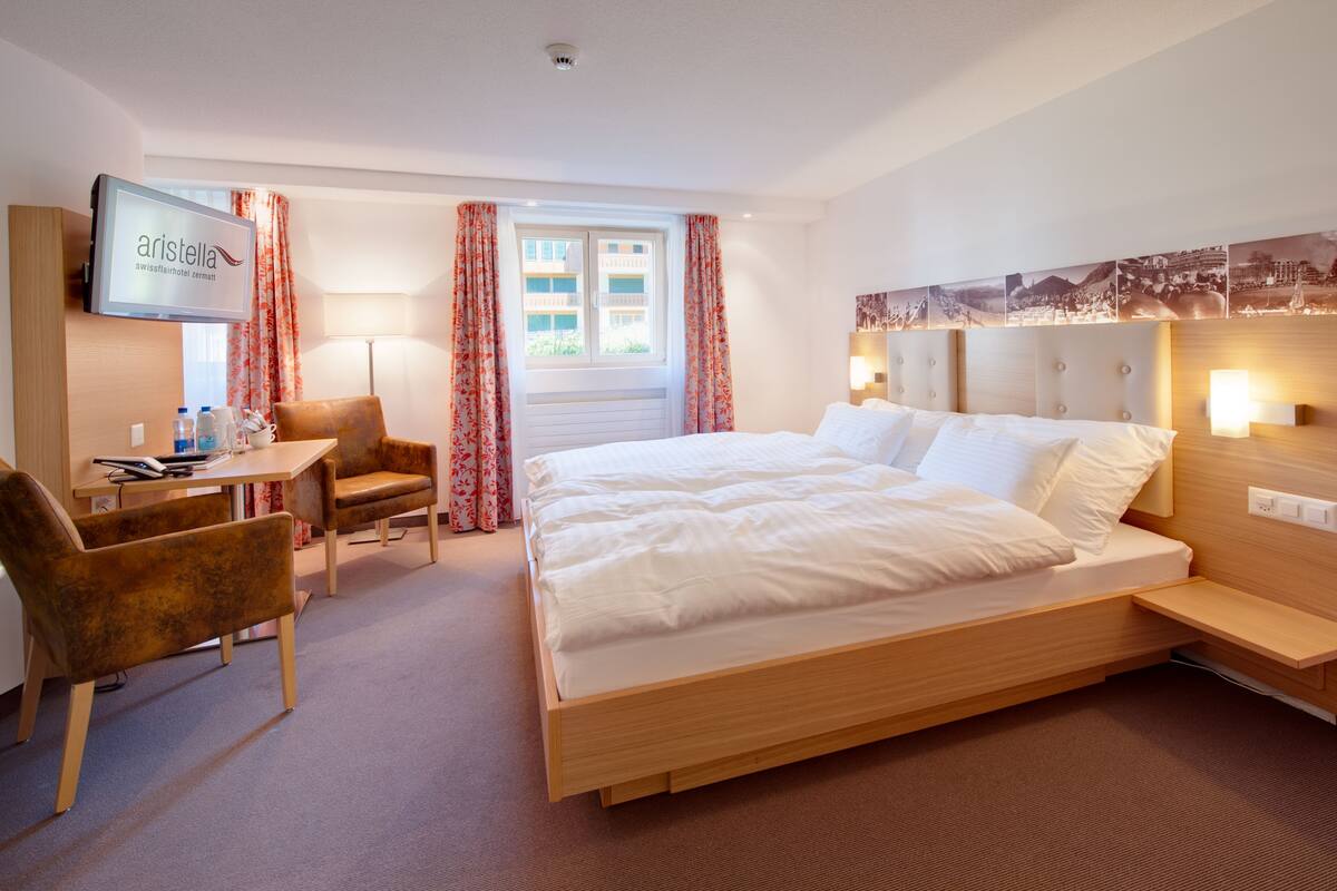 Habitación de hotel con cama, escritorio, silla, televisor y ventana con cortinas.