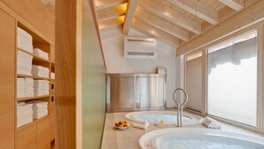 A spa room with two hot tubs, wooden shelving with towels, a fruit bowl, and a large window. - Aristella Swissflair Hotel & Apartements (Zermatt)