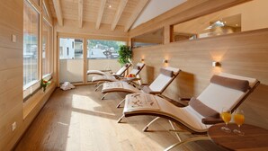 A sunlit room with wooden floors and ceiling, featuring lounge chairs with towels and a table with two glasses of orange juice. - Aristella Swissflair Hotel & Apartements (Zermatt)