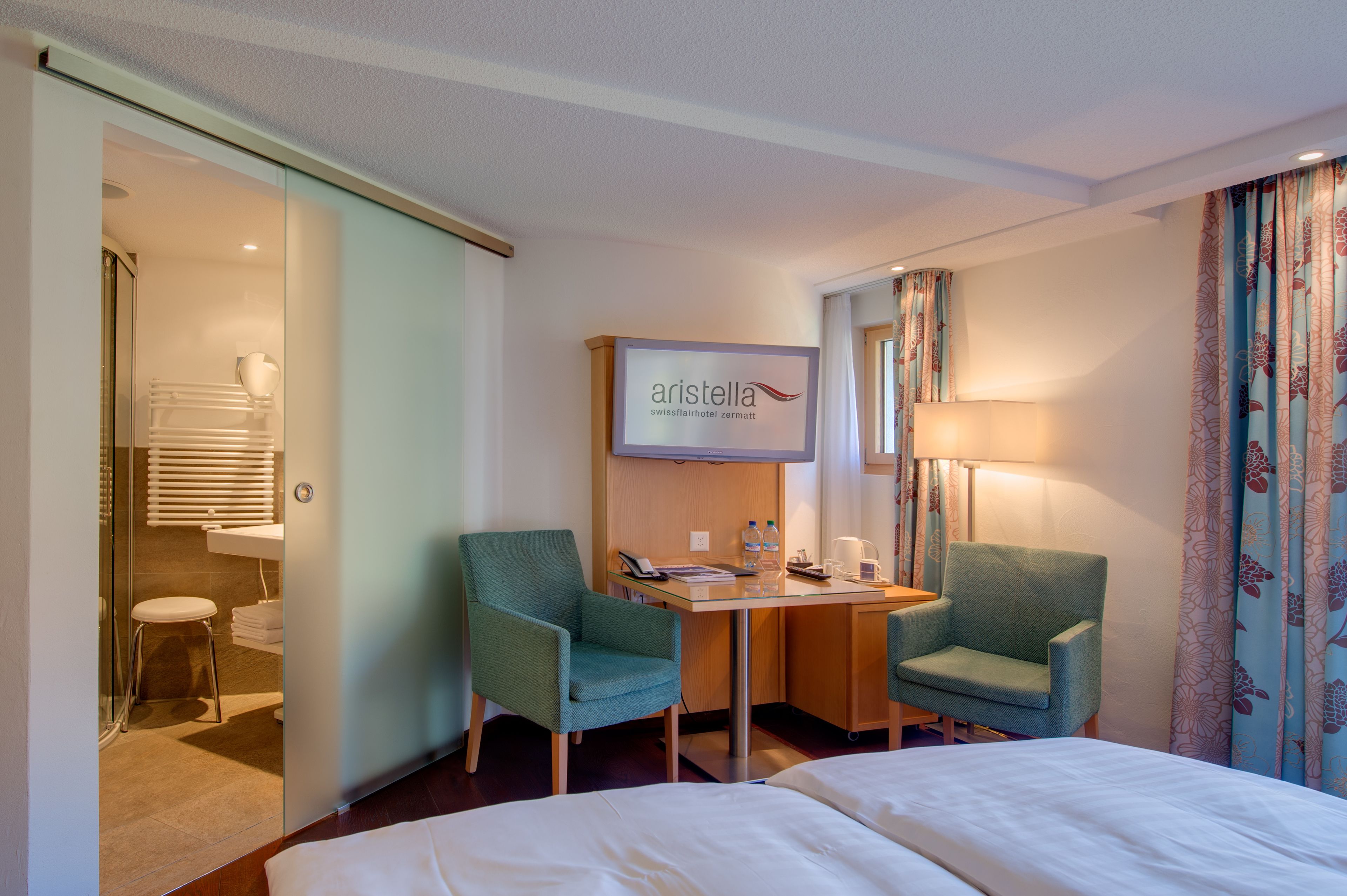 A hotel room with two green armchairs, a glass table with a phone and a lamp, a bed, and a shower area with a towel rack.