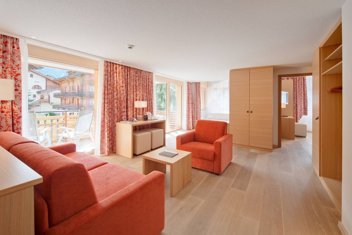 Una habitación de hotel moderna con sofá, sillón y una mesita. Tiene un balcón con vistas a edificios.