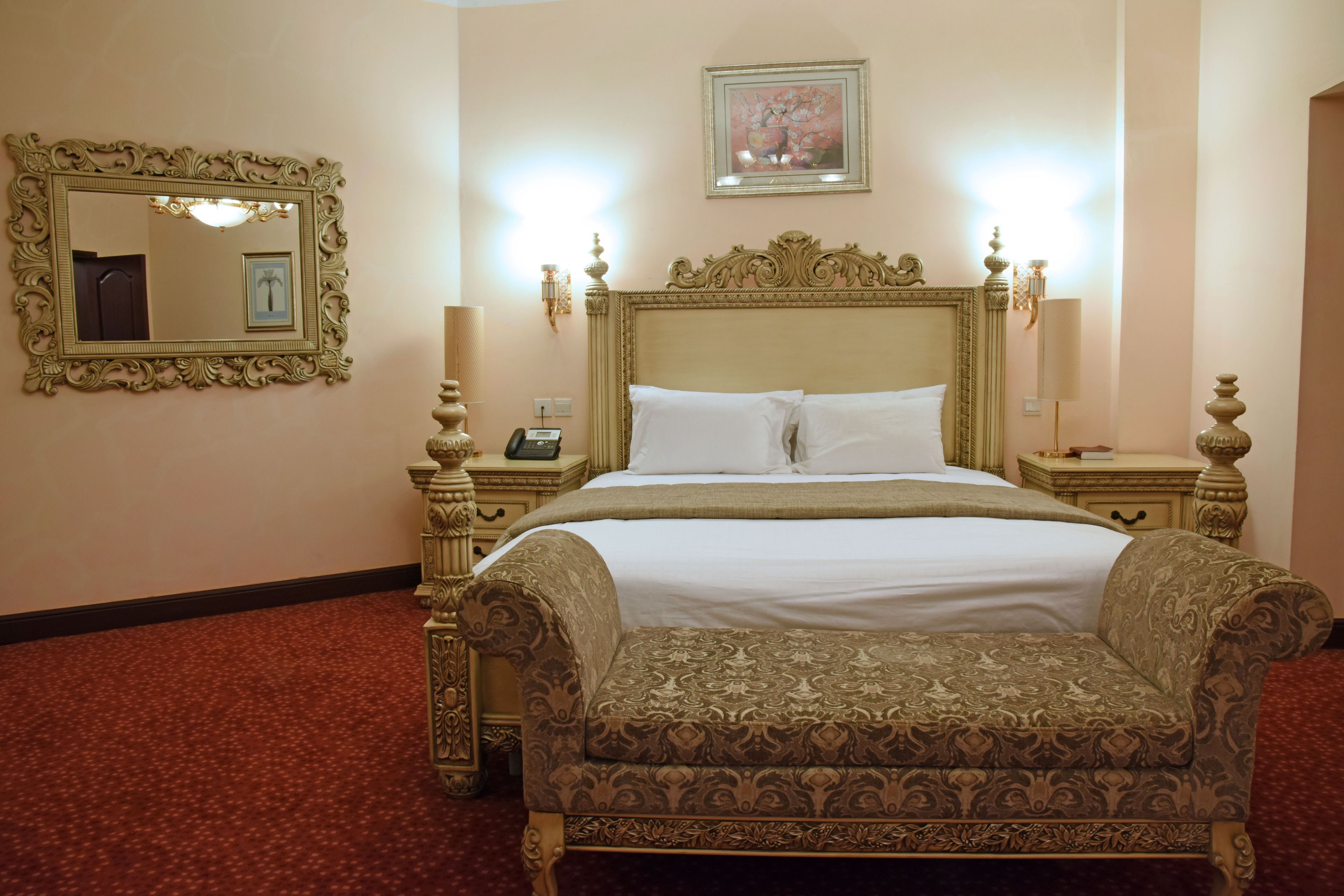 A hotel room with a large bed, ornate headboard, two bedside tables with lamps, a mirror, and a framed picture on the wall.