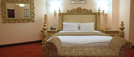 A hotel room with a large bed, ornate headboard, two bedside tables with lamps, a mirror, and a framed picture on the wall.
