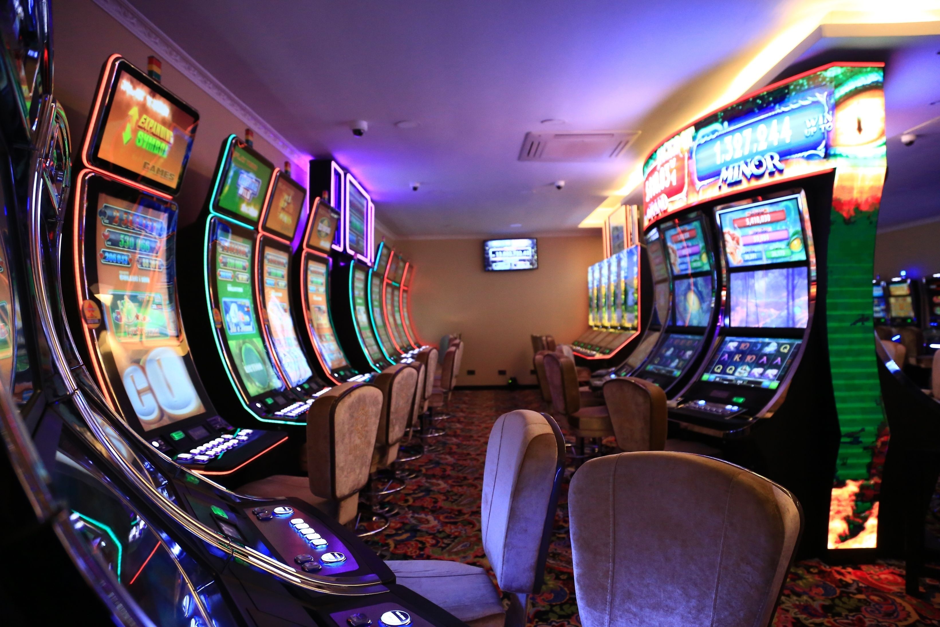 A row of slot machines in a casino with a carpeted floor and chairs in the center.