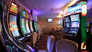 A row of slot machines in a casino with a carpeted floor and chairs in the center.