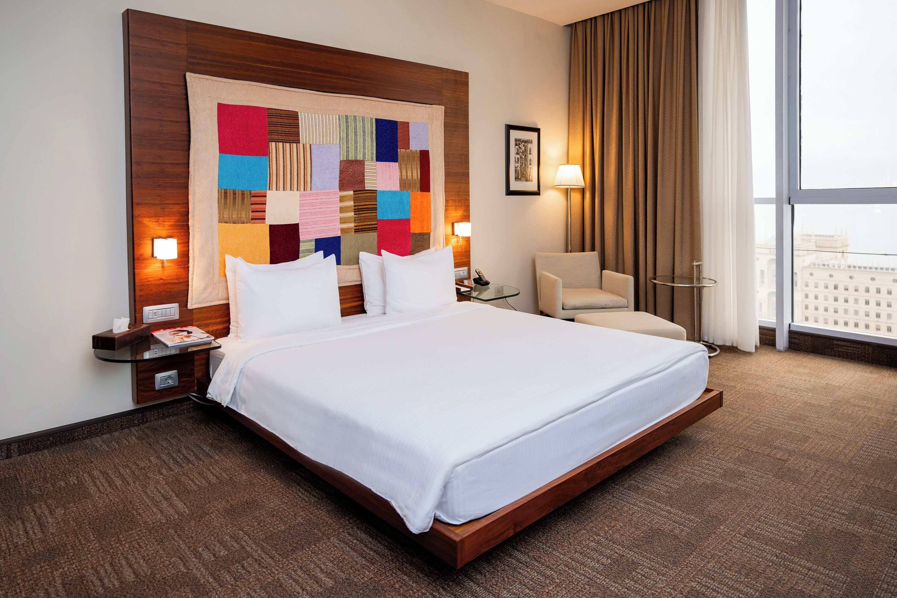 A hotel room with a large bed, a desk, a chair, a nightstand, a lamp, and a view of the city.