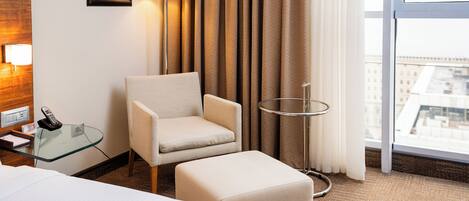 A hotel room with a bed, a chair, a small table with a lamp, a glass table with a phone, and a view of the city through large windows.