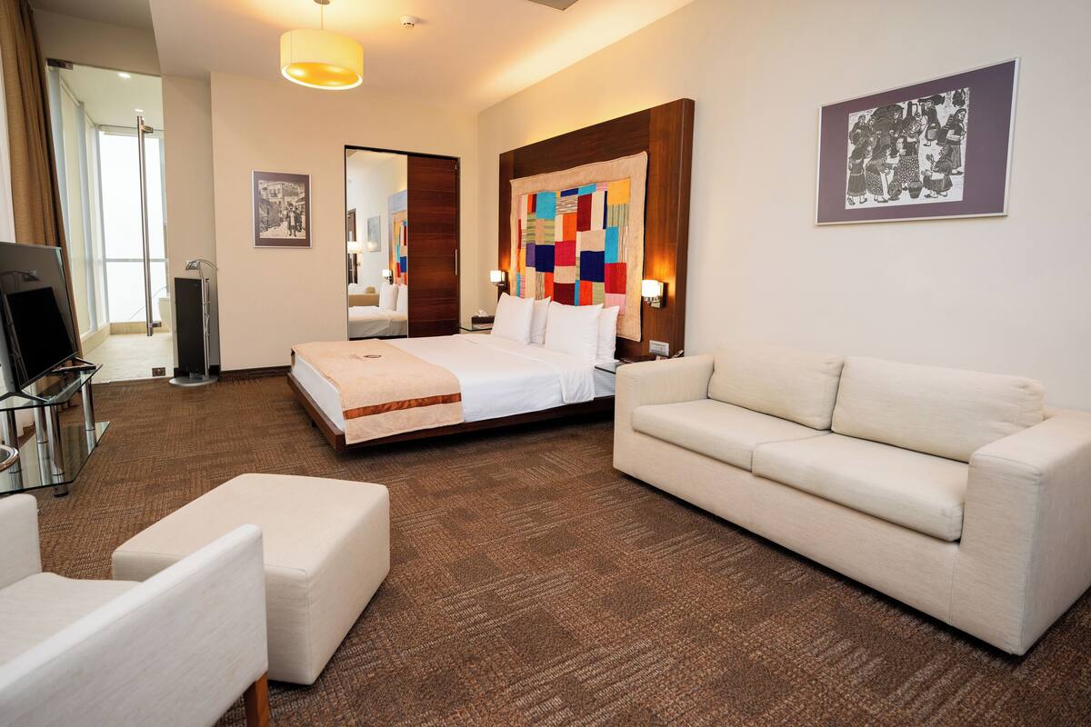 A hotel room with a bed, a sofa, a TV, a mirror, and artwork on the walls.