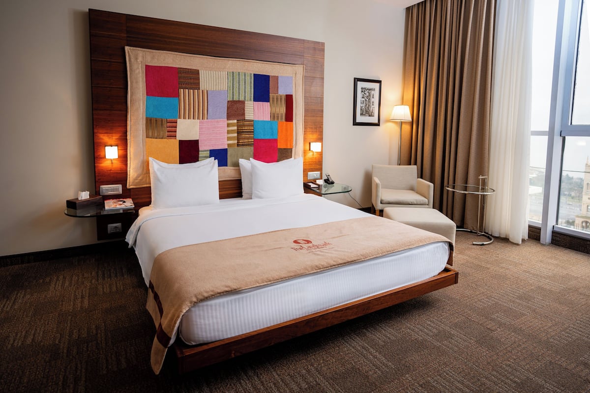 A hotel room with a large bed, a headboard with a colorful quilt, a sitting area with a chair, a small table, and a view through a window.