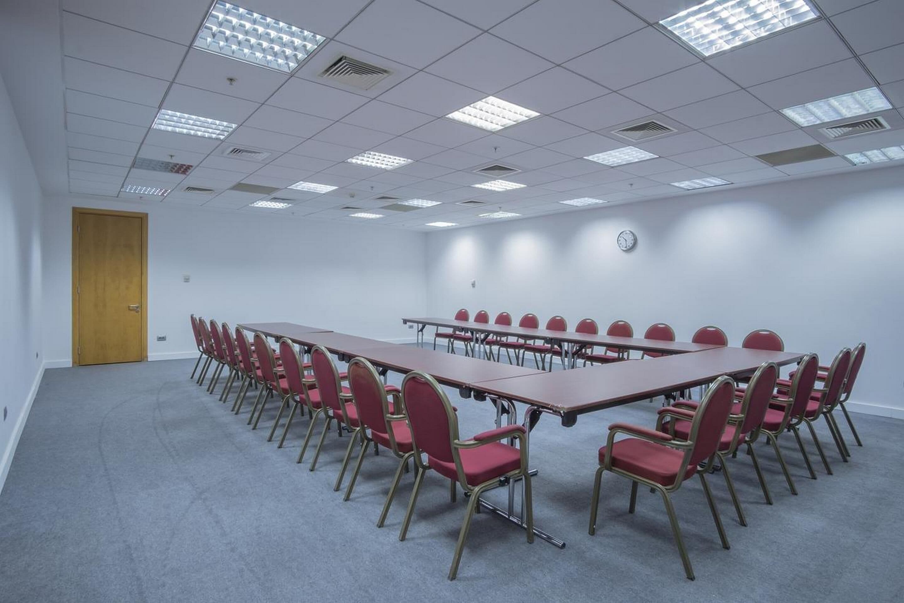 Uma sala de conferências com uma mesa comprida e cadeiras vermelhas, paredes brancas e um relógio na parede.