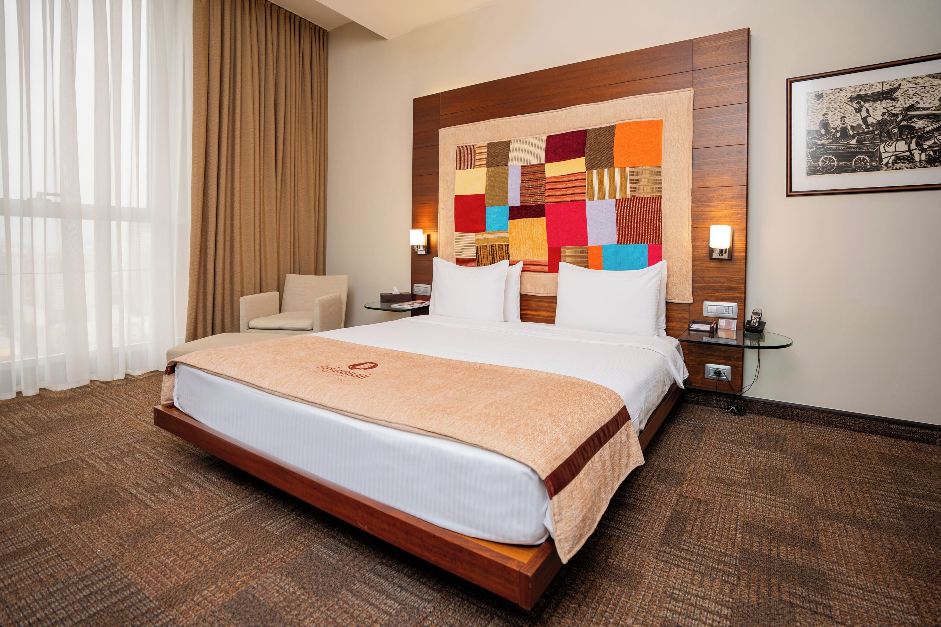 A hotel room with a large bed, a wooden headboard, a colorful quilt, a nightstand, a lamp, and a framed picture on the wall.