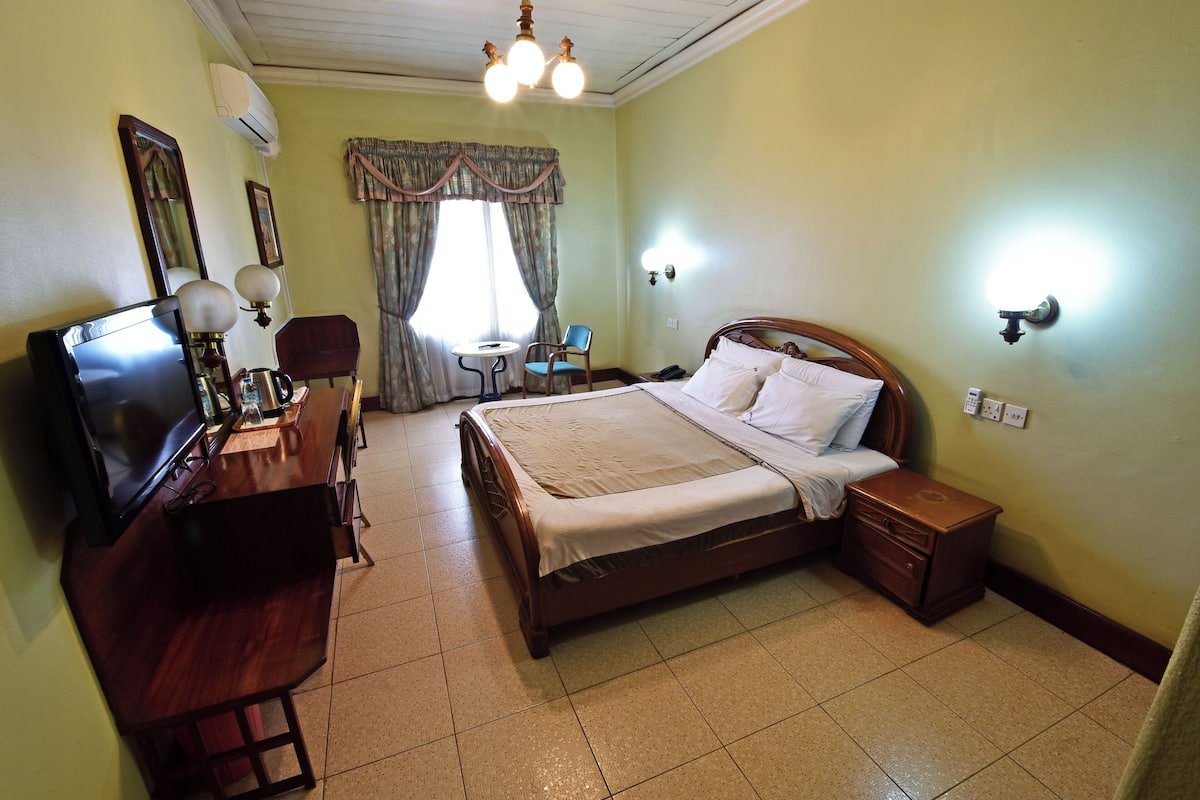 A hotel room with a bed, a desk with a television, a chair, a small table, and a window with curtains.