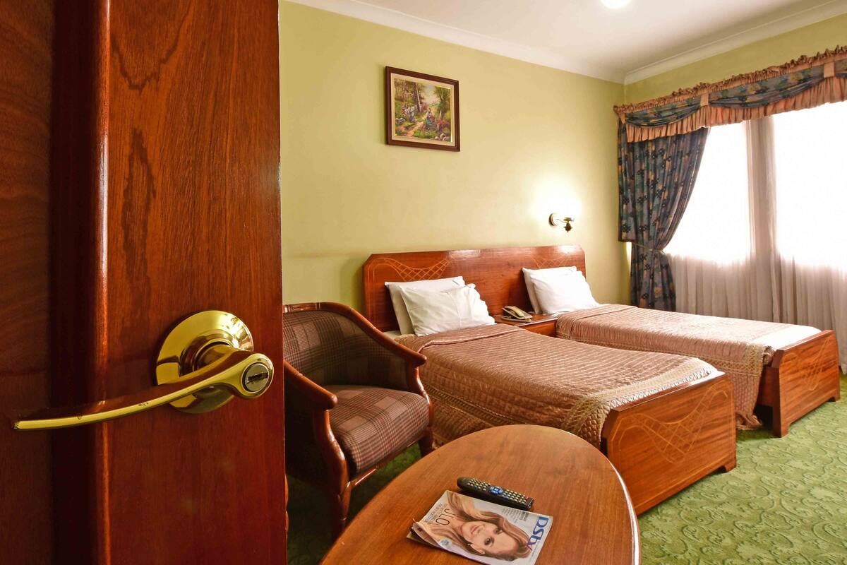 A hotel room with two beds, a wooden chair, a table with a magazine, and a painting on the wall.