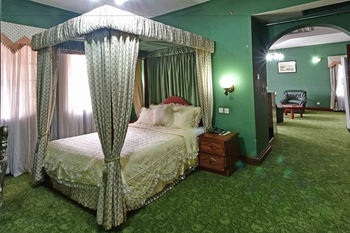 A four-poster bed with a canopy, a nightstand, a chair, and a wall-mounted lamp in a room with green walls and patterned carpet.