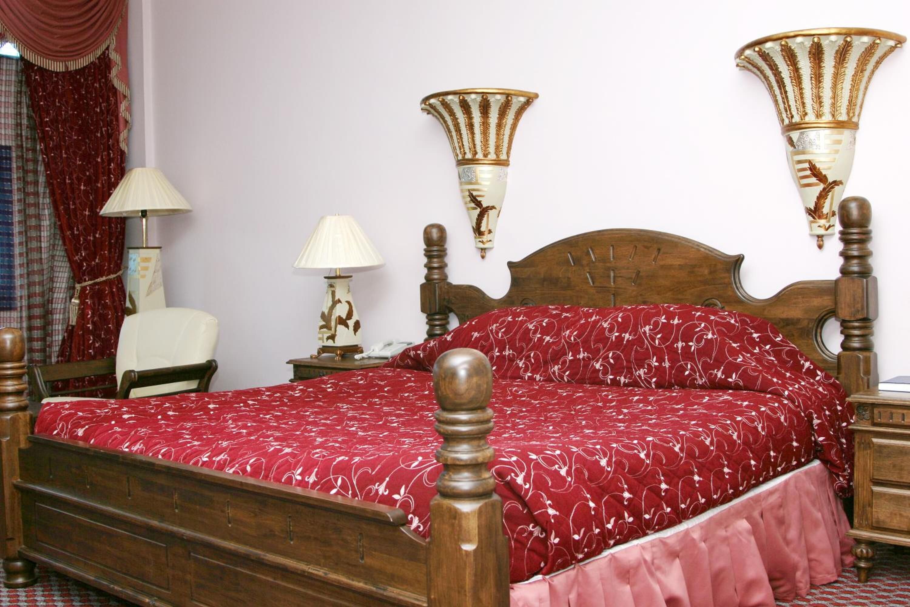 A wooden bed with a red patterned bedspread, two bedside lamps, and a headboard.