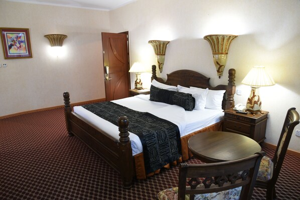 Una cama bien hecha con tablero de madera, dos lámparas de noche, una mesita de noche de madera y una silla con cojín floral.