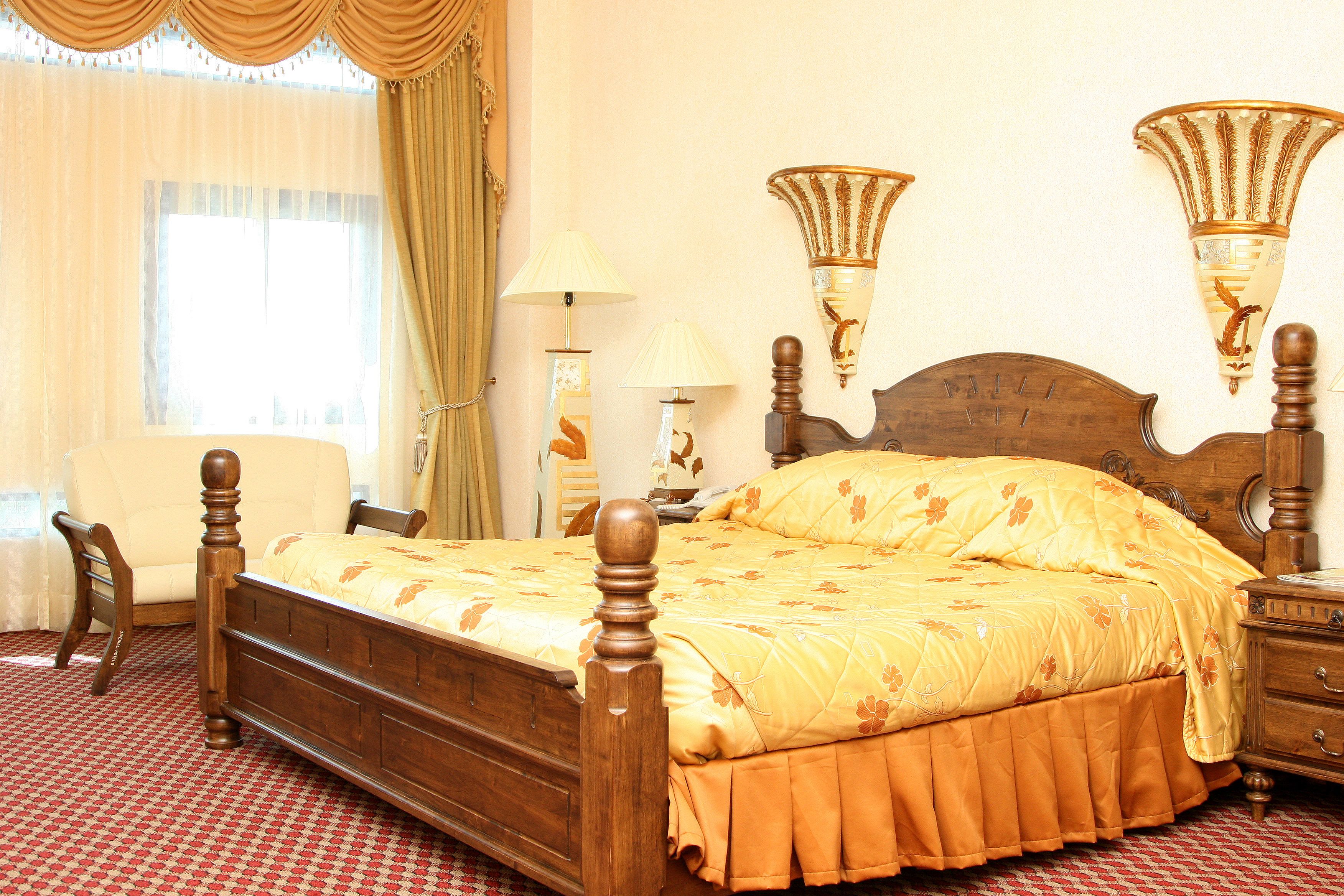 A wooden four-poster bed with a floral bedspread, a wooden nightstand, a chair, and two wall sconces.
