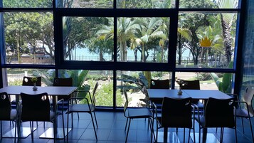 Un restaurante con amplios ventanales que ofrecen vistas a palmeras y a la playa. Las mesas y sillas están dispuestas en el interior.