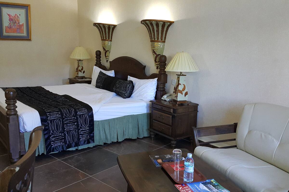 A hotel room with a bed, a nightstand, a lamp, a chair, and a table with a magazine and a bottle.