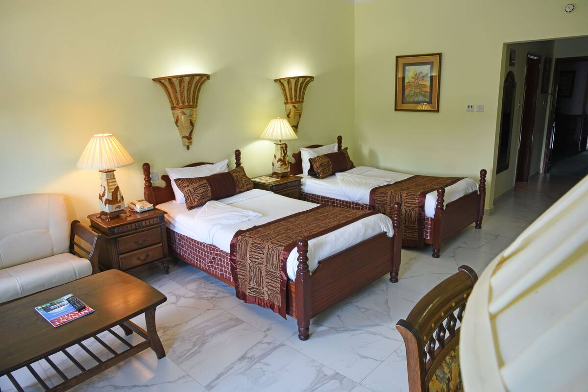 A hotel room with two single beds, a sofa, a wooden table, and wall-mounted lamps.