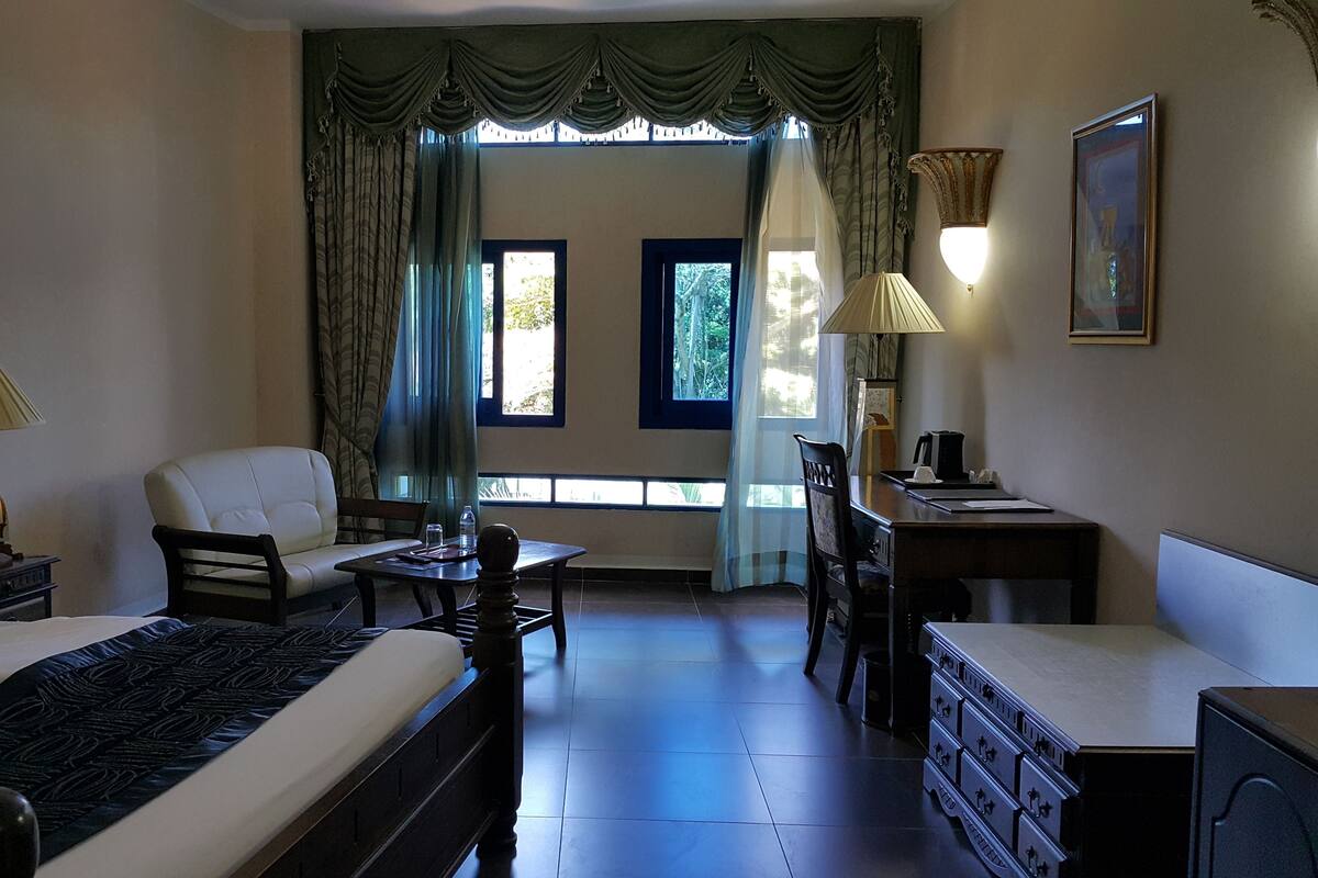 A hotel room with a bed, a chair, a desk, a lamp, a dresser, and a window with curtains.