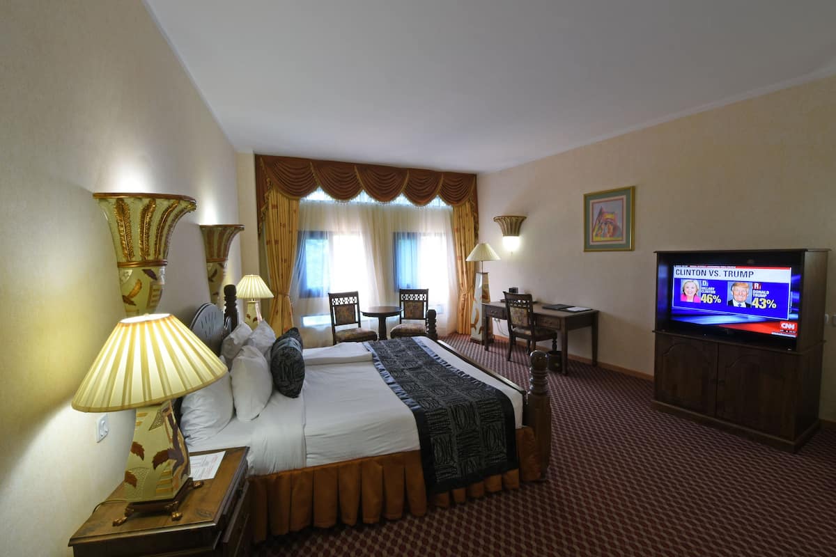 A hotel room with a bed, two lamps, a TV displaying a news channel, a desk, and a chair.