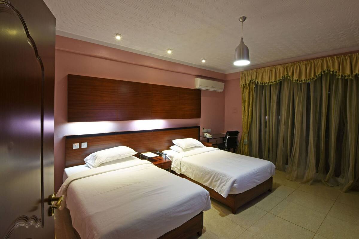 A hotel room with two beds, a wooden headboard, an air conditioning unit, a desk with a chair, and a curtain.