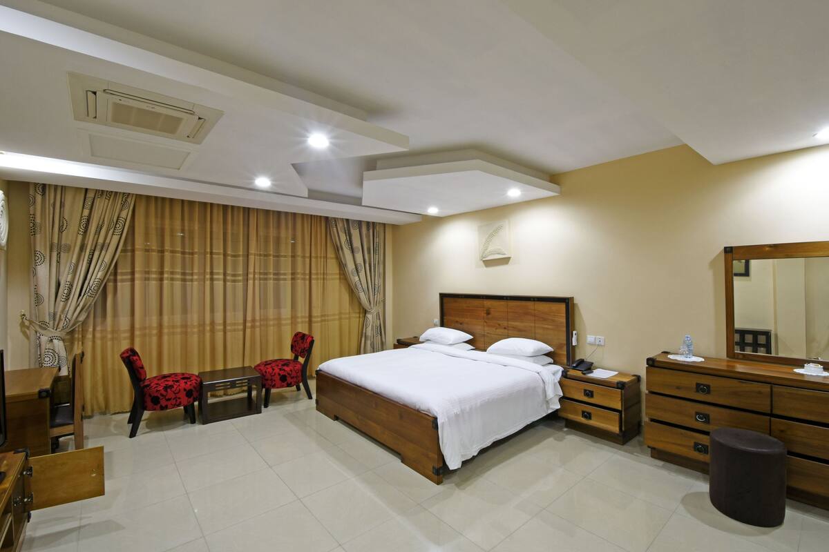 A hotel room with a wooden bed, a wooden dresser, a desk with a chair, and a large mirror.