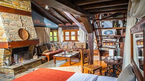 A cozy room with a stone fireplace, wooden beams, a sofa, a coffee table, and a bookshelf. - Oikoperiigitis Hotel (Sintiki)