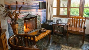 A cozy room with a stone fireplace, wooden furniture, a television mounted on the wall, and a window with a view of greenery outside. - Oikoperiigitis Hotel (Sintiki)