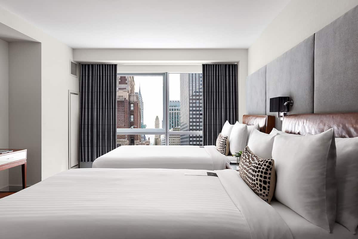 Habitación de hotel con dos camas, un ventanal con cortinas y vistas a la ciudad.