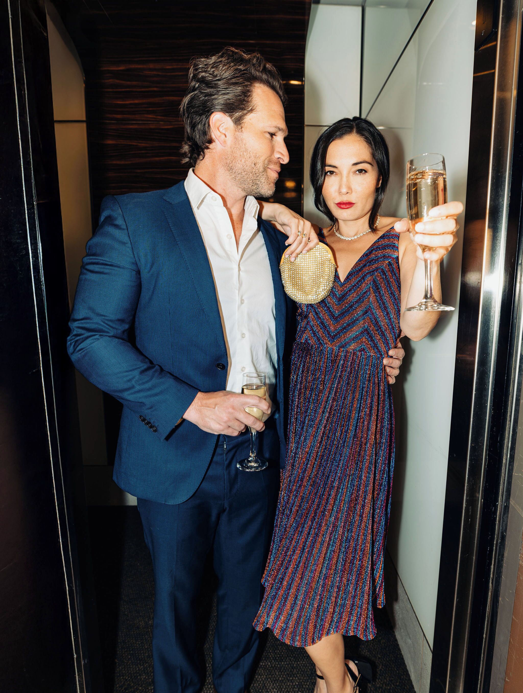 A man and a woman standing in a hallway, holding drinks.