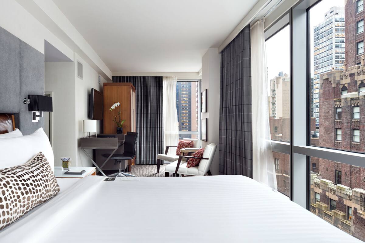Habitación de hotel con una cama grande, un escritorio, una silla y vistas de la ciudad a través de amplios ventanales.