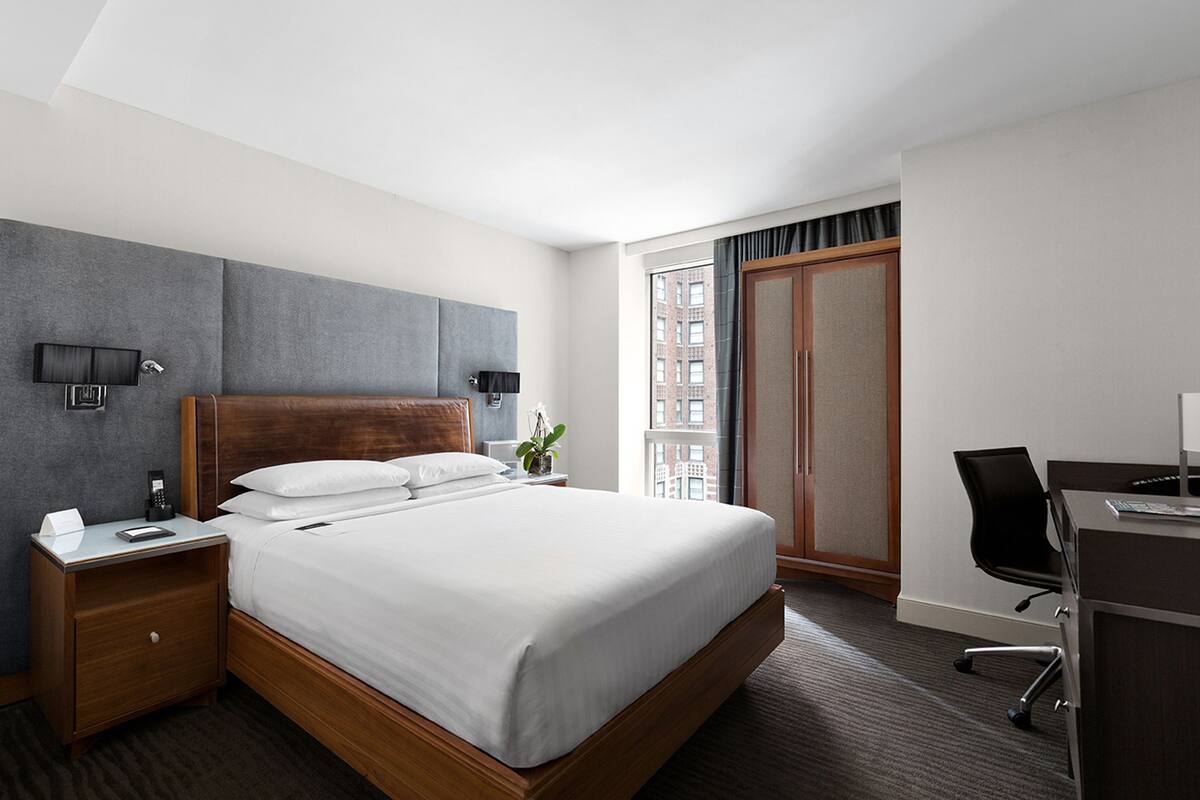 Habitación de hotel con una cama grande, un escritorio, una silla, una mesita de noche, una lámpara y vistas a la ciudad por una ventana.