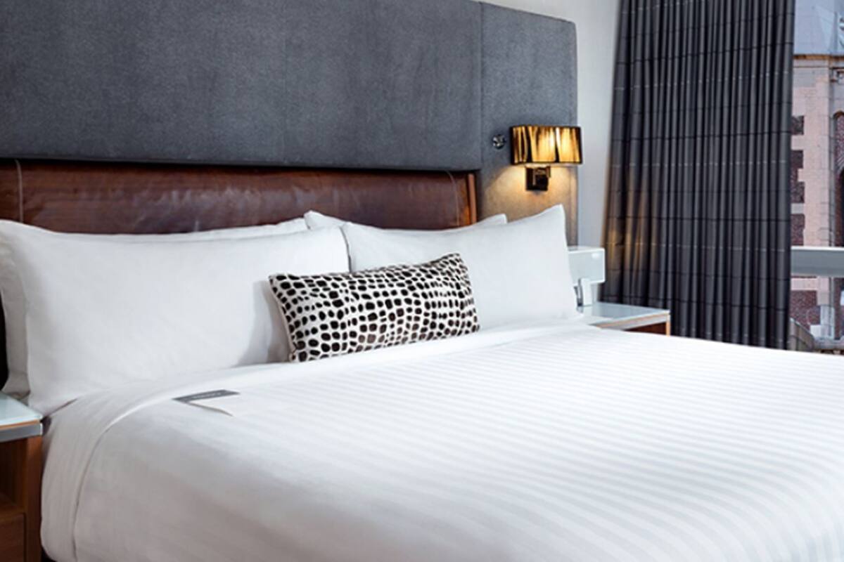 Habitación de hotel con una cama grande, cabecera, mesita de noche, lámpara y ventana con cortinas.