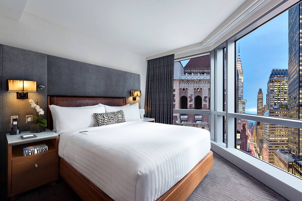 Habitación de hotel con una cama grande, una mesita de noche, una lámpara y vista a la ciudad.