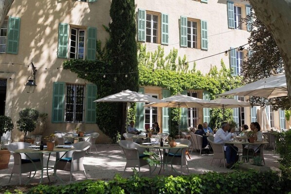 An outdoor café with tables and chairs, surrounded by greenery and a building with shutters. - Relais de la Magdeleine (Gemenos)