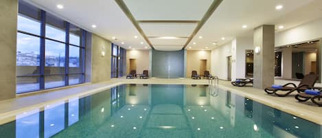 Una piscina interior con agua azul cristalina, tumbonas y ventanas amplias que ofrecen vistas al exterior.