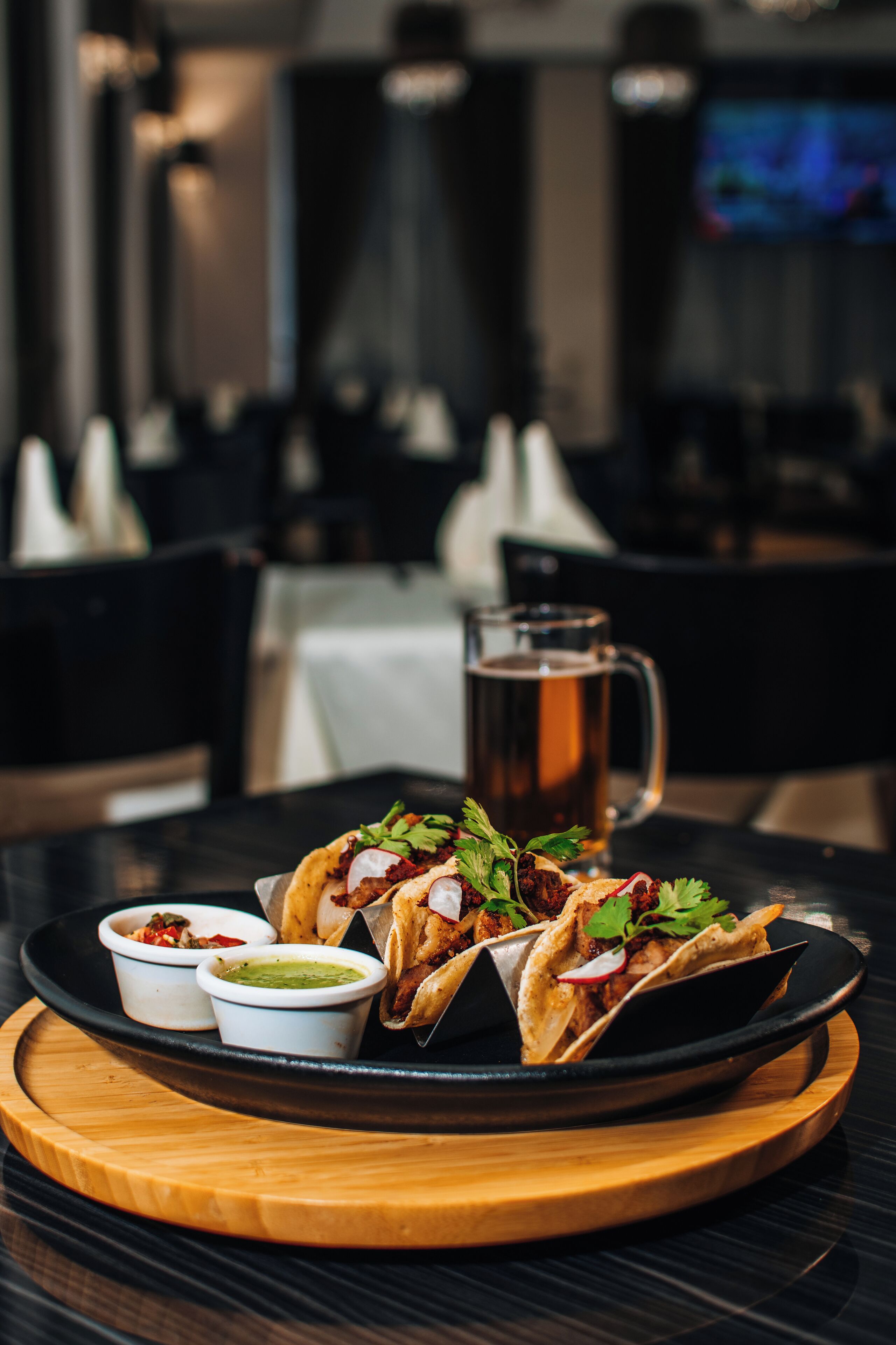 A tray of tacos with condiments on a wooden board, set on a table in a restaurant with a TV in the background.