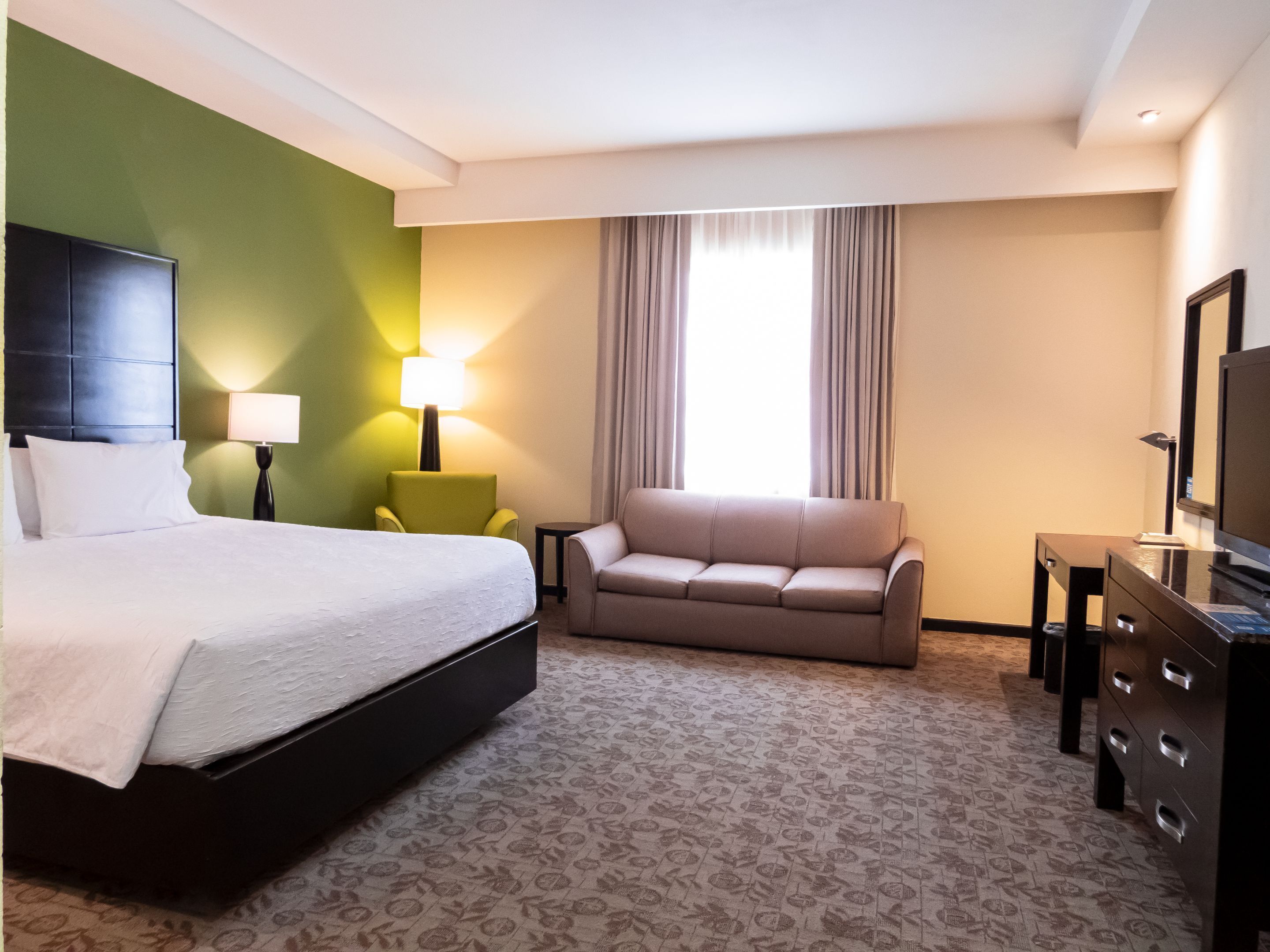 A hotel room with a bed, a sofa, a TV, a lamp, and a window with curtains.