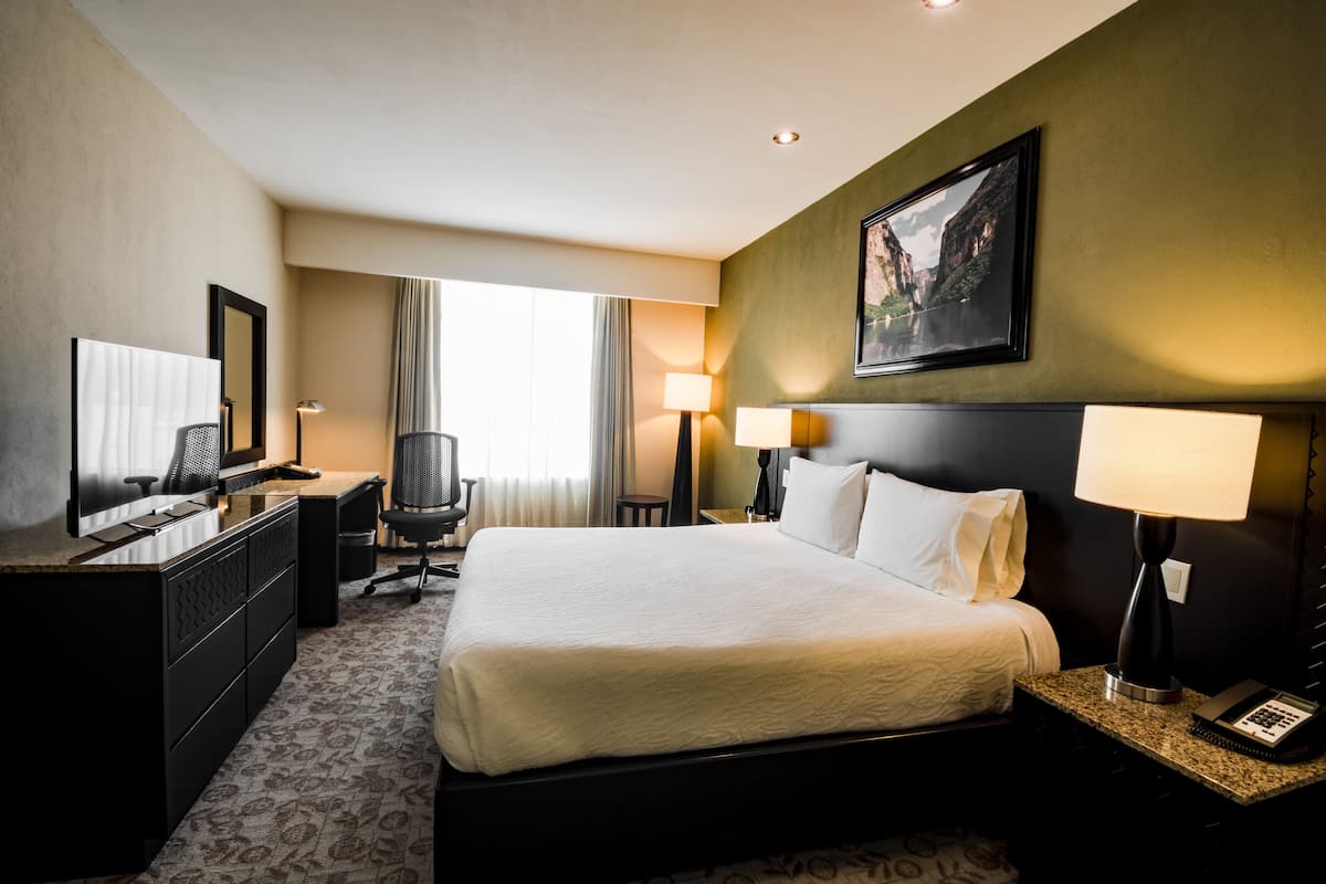 A hotel room with a large bed, a desk with a computer, a chair, a lamp, and a painting on the wall.