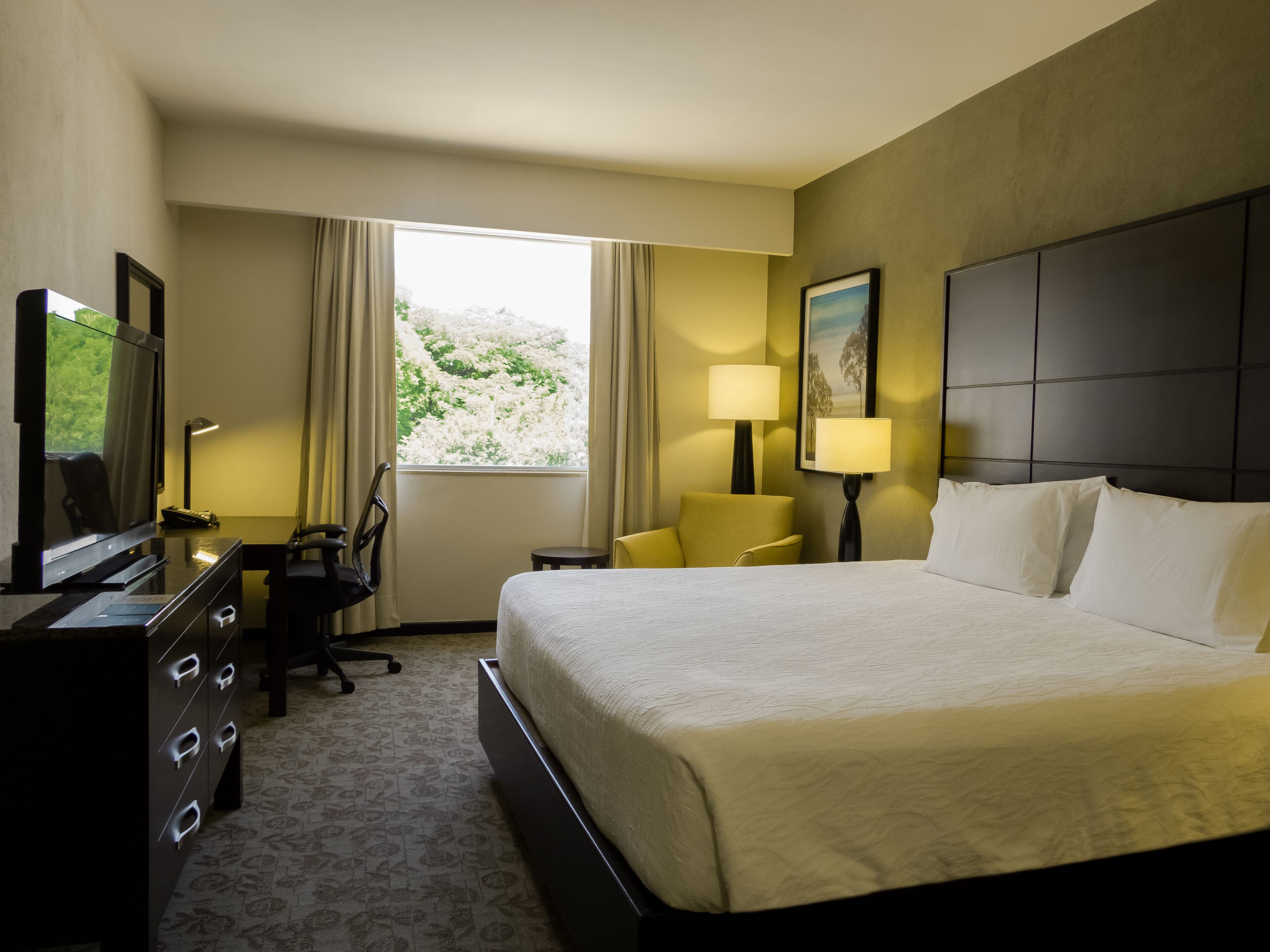 Habitación de hotel con cama, escritorio con silla, televisor, lámpara y ventana con cortinas.